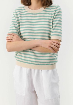 Woman wearing a short-sleeve mint green and cream striped sweater with white loose-fitting pants, arms crossed at the waist.