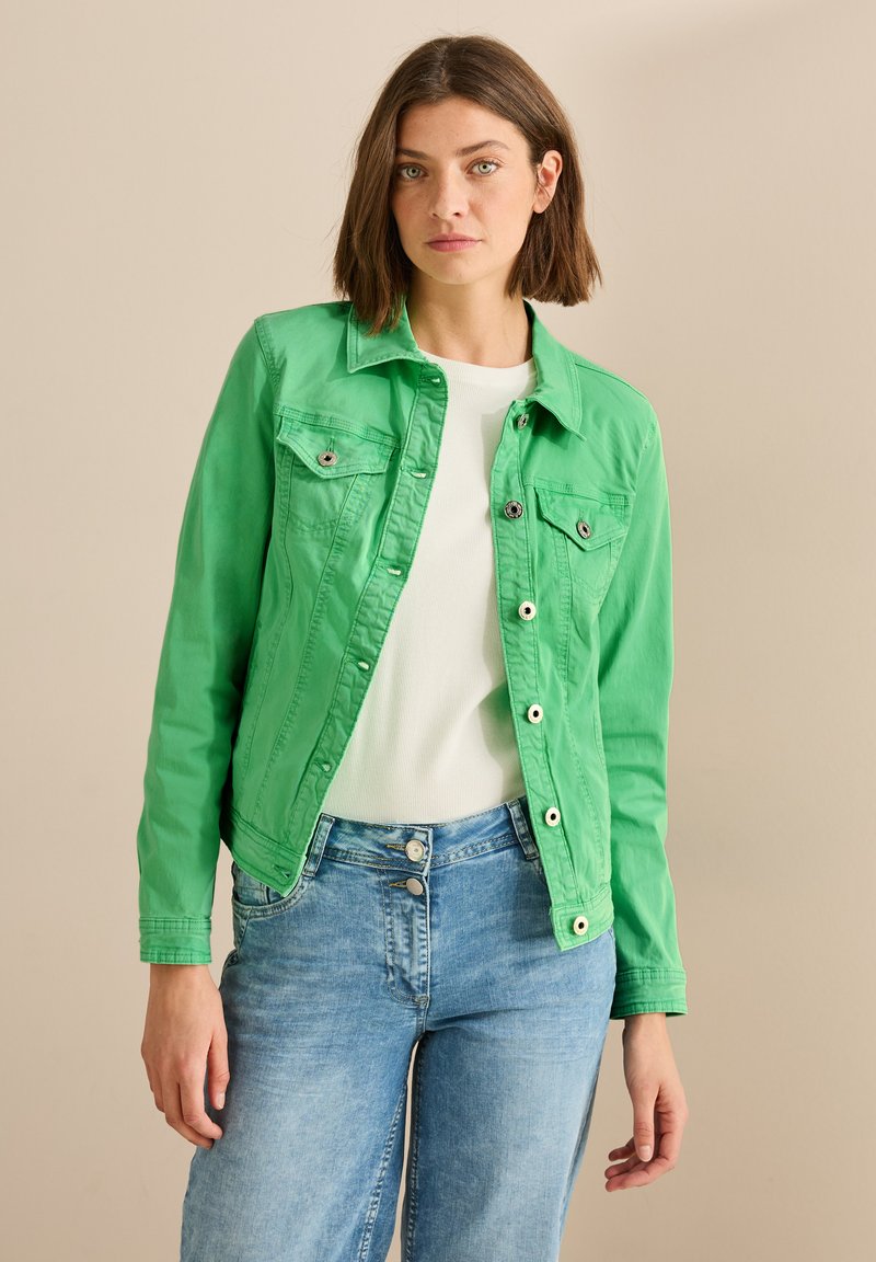 Veste en denim verte, devant boutonné, deux poches poitrine avec rabat, manches longues, design avec col, associée à un haut blanc et un jean bleu clair.