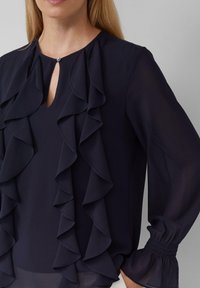 Blusa blu navy con strati di balze lungo il davanti, scollo a chiave e maniche lunghe a palloncino con polsini elasticizzati. Tessuto dalla texture liscia.