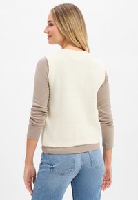 Leichter, cremfarbener Weste mit Rippstruktur und rundem Halsausschnitt, über einem hellbraunen Langarmshirt getragen. Kombiniert mit blauen Jeans.
