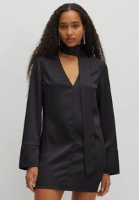 Schwarzes Satin-Kleid mit langen Ärmeln, V-Ausschnitt und geknotetem Bindeband am Hals. Der Stoff hat eine glatte, glänzende Textur.