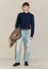 Tânăr îmbrăcat în blugi deschiși la culoare, tricou lung cu mânecă într-o nuanță de albastru închis, curea neagră și pantofi negri, ținând în mână o geacă de piele maro pliată.