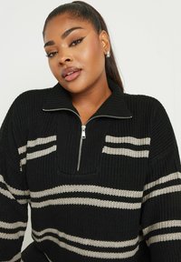 Pull en tricot noir avec un col zippé mi-haut, présentant des rayures horizontales beiges et un motif texturé sur l'ensemble.