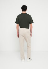 Beige Jogginghose mit einer Gesäßtasche, kombiniert mit einem dunkelgrünen T-Shirt. Das Outfit zeichnet sich durch eine lockere Passform und einen elastischen Bund aus. Weiße Sneakers.