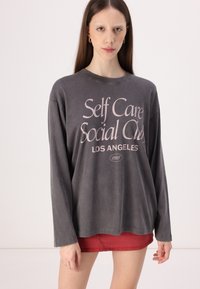Grå langermet skjorte med et falmet rosa grafisk tekst "Self Care Social Club LOS ANGELES 1995" på forsiden. Myk stoff, avslappet passform.