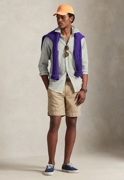 Μοντέλο που φοράει ριγέ πουκάμισο με κουμπιά, χακί σορτς, μωβ πουλόβερ περασμένο στους ώμους, πορτοκαλί καπέλο και μπλε αθλητικά παπούτσια.