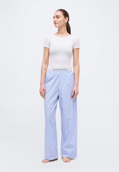 Γυναίκα όρθια, ξυπόλυτη, φοράει εφαρμοστό λευκό κοντομάνικο τοπ και φαρδιά μπλε-λευκή παντελόνα με κάθετες ρίγες και ζώνη με λογότυπο.