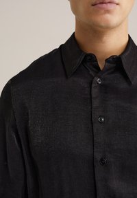 Mees, kes kannab musta tekstureeritud nööpidega särki terava kraega, nähtav kaelast kuni ülemise rindkere osani neutraalse tausta ees.