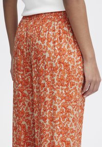 Kalhoty s květinovým potiskem v oranžových tónech na krémovém podkladu. Měkká látka, elastický pas a volný střih, který zdůrazňuje strukturovaný vzor.