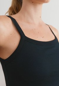 Débardeur noir à côtes avec fines bretelles et décolleté carré. Présente une texture due aux côtes et un design ajusté. Gros plan sur le haut du corps.