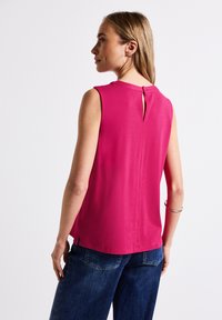 Top senza maniche di un vivace colore rosa, con chiusura a chiave sul retro, texture liscia e spacchi laterali per maggiore comfort. Abbinato a pantaloni in denim blu.