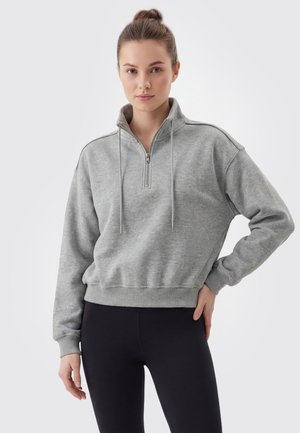 Ung kvinde med håret i en knold bærer en grå trøje med kvart lynlås og sorte leggings mod en ensfarvet lys baggrund.