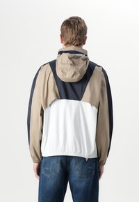 Fargeblokkert vindjakke med et design i beige, marineblå og hvit. Lett stoff med justerbar hette og elastisk kant.