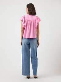 Femme se tenant de dos, portant un haut rose ample à volants à manches courtes et un jean large bicolore bleu avec des chaussures à talons noires.