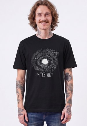Jeune homme avec des tatouages souriant, portant un t-shirt noir avec un design blanc de la galaxie de la Voie lactée et un texte sur le devant.