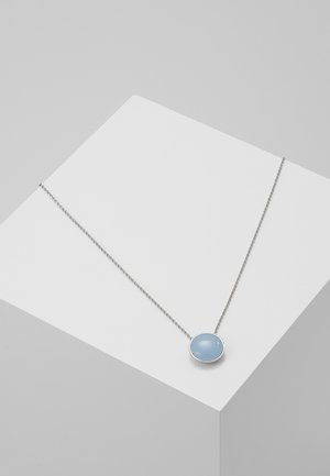 Collier en chaîne argentée avec un pendentif rond bleu clair reposant sur un socle géométrique blanc sur un fond gris.