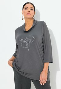 Top gris con cuello de capucha y mangas de tres cuartos, que presenta un diseño floral bordado en blanco. Confeccionado en una tela suave y flexible.