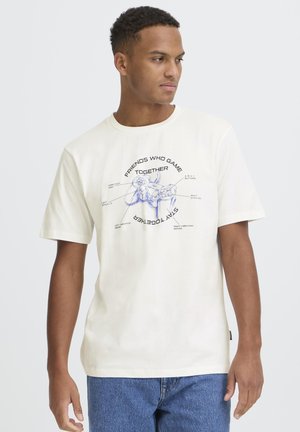 Hombre con camiseta blanca con un dibujo azul de un mando de juego y botones etiquetados, combinado con jeans azules, mirando hacia su derecha.