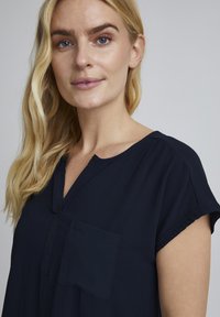 Tamsiai mėlyna trumpa rankovė viršutinė dalis su v-švarku ir mažomis priekine užtrauktuku. Audinys atrodo minkštas, su lygiu tekstūra ir minimaliais siuvimo detalėmis.