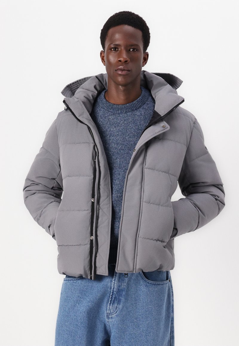 Joven vestido con una chaqueta acolchada gris sobre un suéter de punto azul y jeans sueltos de un azul claro, de pie con las manos en los bolsillos frente a un fondo blanco.