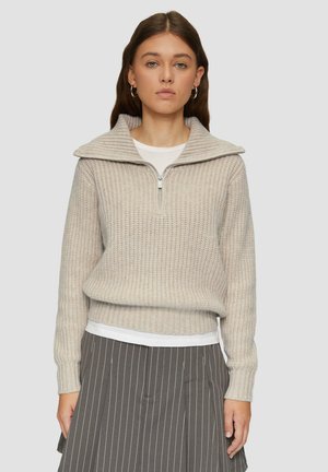 Light beige knit sweater with a zippered collar, ribbed texture, and long sleeves, worn over a white shirt and paired with a striped skirt.