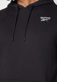 Sweat à capuche noir Reebok avec une poche frontale, des cordons de serrage et un logo blanc sur la poitrine. Tissu doux, design classique et style à capuche.