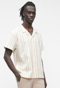 Mężczyzna z dredami, ubrany w kremową koszulę z cienkimi pionowymi paskami i beżowe spodnie, stoi z jedną ręką w kieszeni na białym tle.