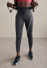 Szare legginsy o wysokim stanie o gładkiej fakturze, zestawione z czarnymi butami sportowymi, trzymające opaski oporowe z czarnymi uchwytami.