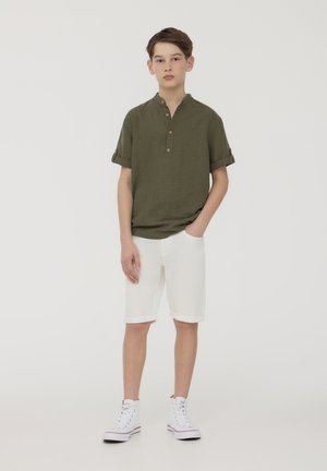 Camicetta verde oliva a maniche corte con colletto alla mandarino e dettagli con bottoni, abbinata a pantaloni corti bianchi con orlo arrotolato e sneaker alte bianche.