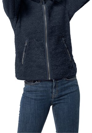 Personne portant une veste polaire zippée bleu foncé et un jean bleu, les bras levés au-dessus de la tête, cadrée à mi-torse.
