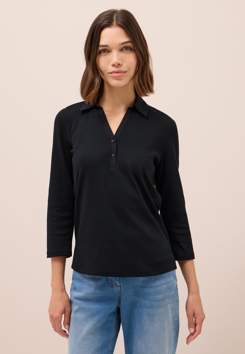 Polo-shirt noir à manches longues avec col, trois boutons à l'avant, en tissu lisse, associé à un jean en denim bleu clair.