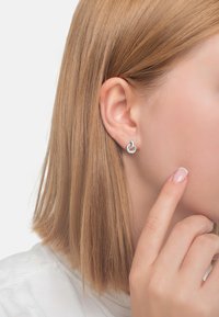 Silverfärgade cirkulära örhängen med en stavdetalj och en slät yta. Modellen håller sitt finger mot kinden och visar upp örhängena.