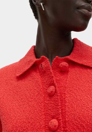 Donkerhuidige persoon draagt een felrood, gestructureerd knoopjasje met kraag en een zilveren oorring in de vorm van een ring.