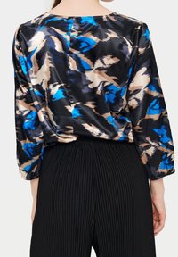 Satinblus med svart bas som har blå och beige abstrakta mönster; långärmade med elastiska manschetter; kombinerad med svarta ribbade byxor.