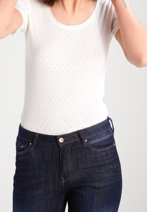 Kobieta ubrana w dopasowany biały krótki top i ciemnoniebieskie dżinsy z wysokim stanem na jednolitym tle.