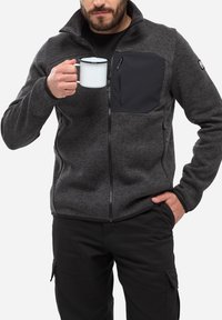 Veste en polaire grise avec fermeture éclair à l'avant, col haut et poche poitrine noire. Le modèle tient une tasse blanche, portant un pantalon noir.