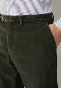 Temnozeleni kordurojski hlače z navpičnimi rebri, z gumbom za zapiranje in stranskimi žepi, v kombinaciji z svetlo modro črtasto srajco.
