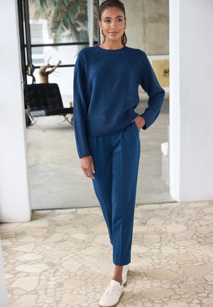 Blauer Strickpullover mit gerippter Struktur, kombiniert mit blauen, schmal zulaufenden Hosen und weißen Sneakers. Lässiges Outfit mit entspanntem Sitz.