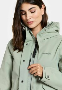 Vaaleanvihreä vedenpitävä takki hupulla, vetoketjulla etuosa ja nepparit. Takissa on mustia yksityiskohtia ja pieni logo rinnassa.