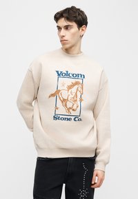 Νεαρός άνδρας που φοράει μπεζ φούτερ Volcom Stone Co. με γραφικό σχέδιο αλόγου και αστεριών, σε συνδυασμό με μαύρο παντελόνι με σχέδιο με τρουκς.