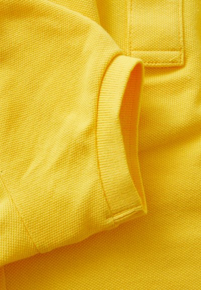 Tessuto giallo brillante con texture a coste e dettagli di cucitura. Primo piano della manica e del corpo di un indumento, che mette in mostra la qualità del materiale.