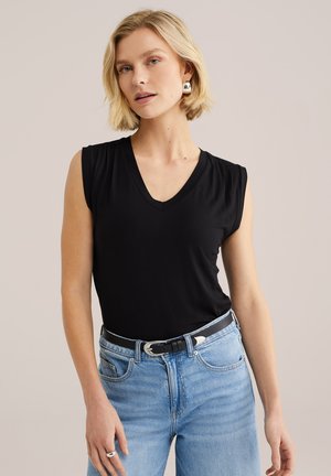 Donna bionda che indossa un top nero senza maniche con scollo a V, jeans azzurri con cintura nera, e orecchini e anello argentati.
