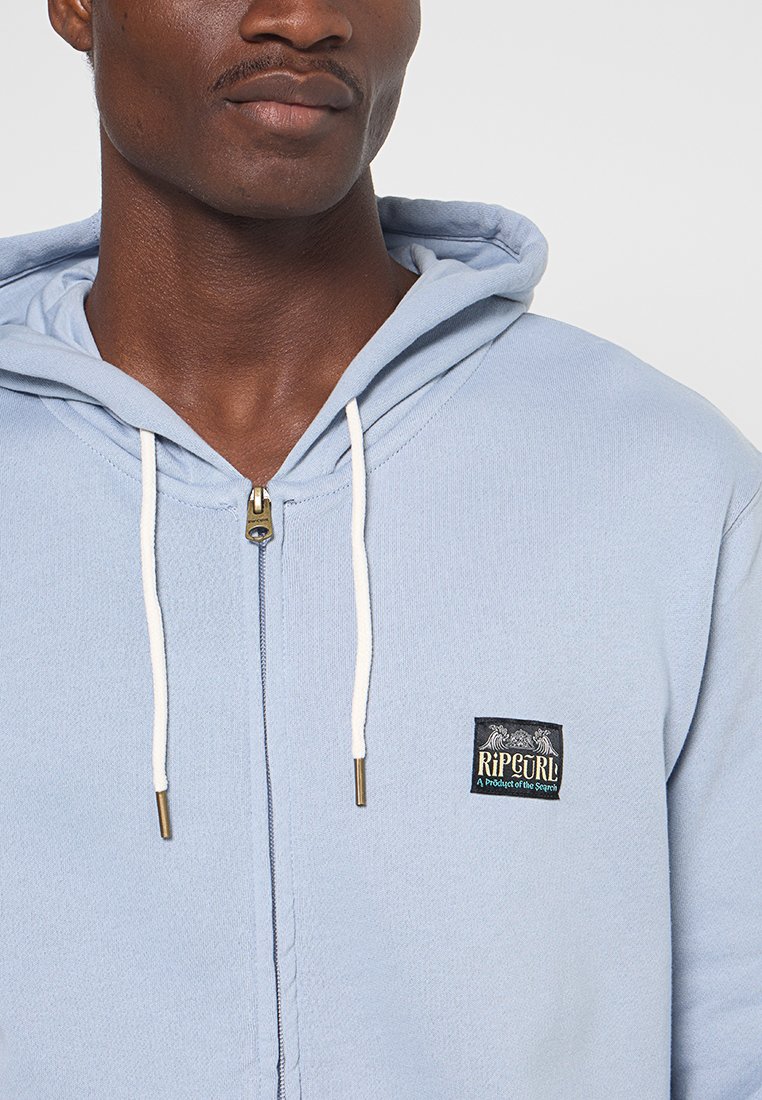 Hellblaues Zip-Hoodie aus weichem Stoff. Verfügt über eine weiße Zug-Schnur, einen goldenen Reißverschluss und ein Logopatch auf der linken Brustseite.
