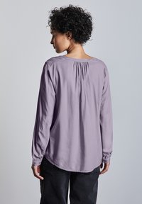 Lila långärmad blus med rund halsringning, rynkdetaljer på baksidan och elastiska ärmslut, bärs med svarta byxor. Slät tygstruktur.