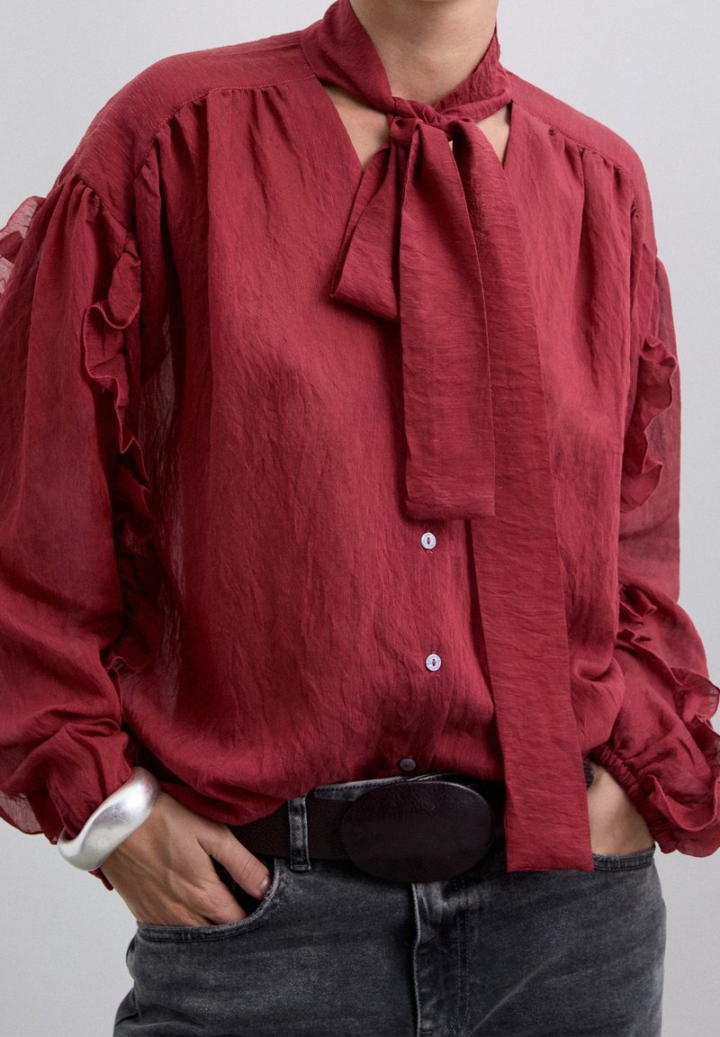 Personne portant un chemisier rouge ample avec des manches à volants et un foulard noué autour du cou, rentré dans un jean noir délavé avec une grande boucle de ceinture.
