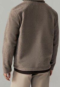 Braune Fleecejacke mit einer strukturierten, plüschigen Oberfläche und einem Kragen. Verfügt über lange Ärmel und einen geraden Saum, getragen über hellen Hosen.