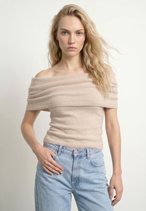 Młoda kobieta z długimi blond włosami, ubrana w beżową bluzkę odkrywającą ramiona i jasnoniebieskie dżinsy na jednolitym białym tle.