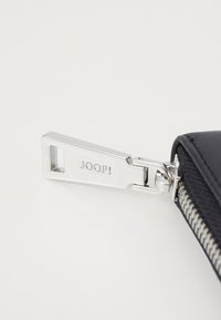 Černý zipový stahovák se stříbrným kováním, na kterém je vyryto "JOOP!". Stahovák má obdélníkový tvar a hladký povrch.