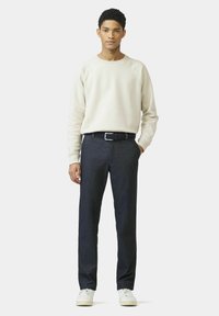 Junger Mann, der ein cremefarbenes Sweatshirt, dunkelblaue Hose mit Gürtel und weiße Sneaker vor einem hellen, einfarbigen Hintergrund trägt.