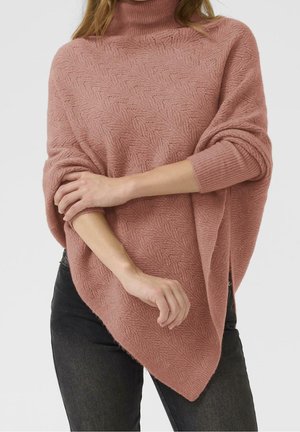 Kvinde iført en tekstureret støvet lyserød turtleneck ponchosweater med flagermusærmer og sorte bukser, poserer med armene krydset.